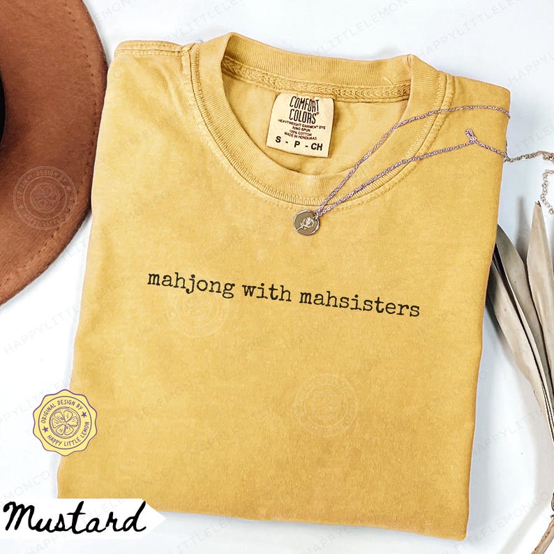 May include: Mustard-colored t-shirt with the text "mahjong with mahsisters" in black. The shirt has a crew neck and short sleeves. A silver necklace with a small pendant is on the shirt. The shirt is labeled "Comfort Colors".