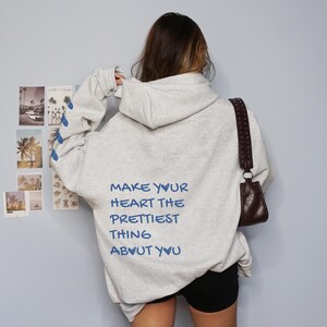 May include: A light gray hoodie with the text "MAKE YOUR HEART THE PRETTIEST THING ABOUT YOU" in blue. The hoodie has a drawstring hood and long sleeves with blue pineapple designs. A brown purse hangs from the shoulder.