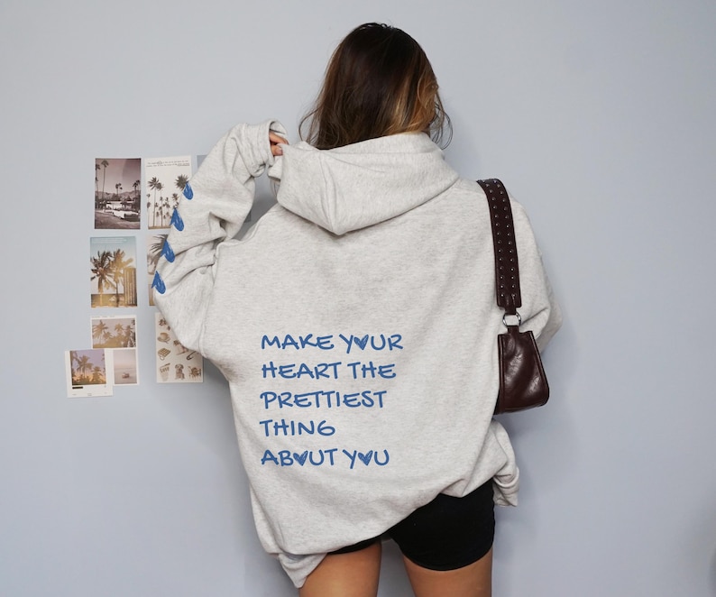 May include: A light gray hoodie with the text "MAKE YOUR HEART THE PRETTIEST THING ABOUT YOU" in blue. The hoodie has a drawstring hood and long sleeves with blue pineapple designs. A brown purse hangs from the shoulder.