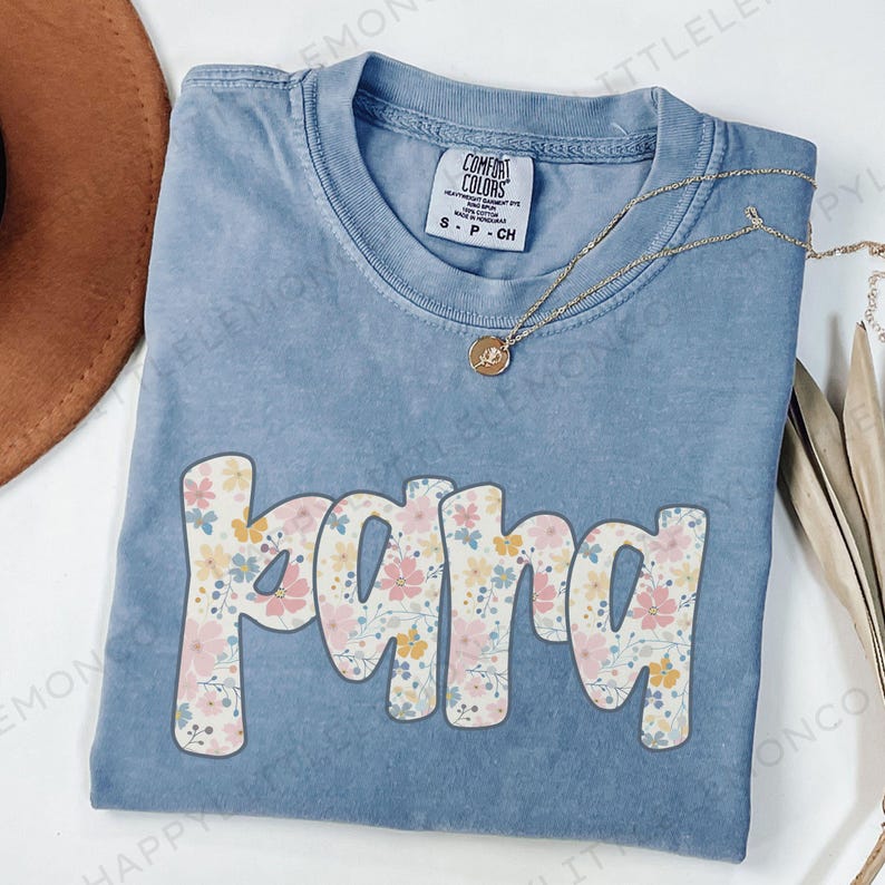 May include: A blue Comfort Colors t-shirt with the word "mama" in a floral design. The shirt has a crew neck and short sleeves. A gold necklace with a pendant is draped across the shirt.