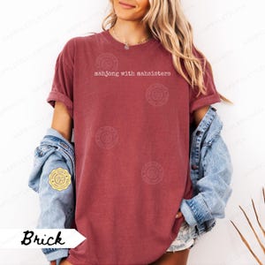 May include: Brick-colored t-shirt with the text "mahjong with mahsisters" in white. The shirt has rolled-up sleeves and is worn with a denim jacket. The shirt is a casual style.