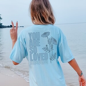 May include: Light blue t-shirt with the text "YOU ARE SO LOVED" in a vertical layout, accompanied by a floral design. The back of the shirt also features the Bible verse John 3:16. The shirt is a soft, comfortable material.