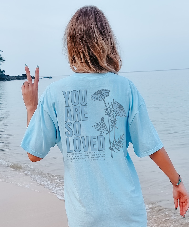 May include: Light blue t-shirt with the text "YOU ARE SO LOVED" in a vertical layout, accompanied by a floral design. The back of the shirt also features the Bible verse John 3:16. The shirt is a soft, comfortable material.