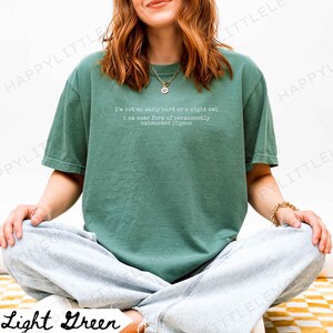 Puede incluir: Camiseta verde claro con el texto "I'm not an early bird or a night owl, I am some form of permanently exhausted pigeon." La camiseta es de color liso y tiene mangas cortas.