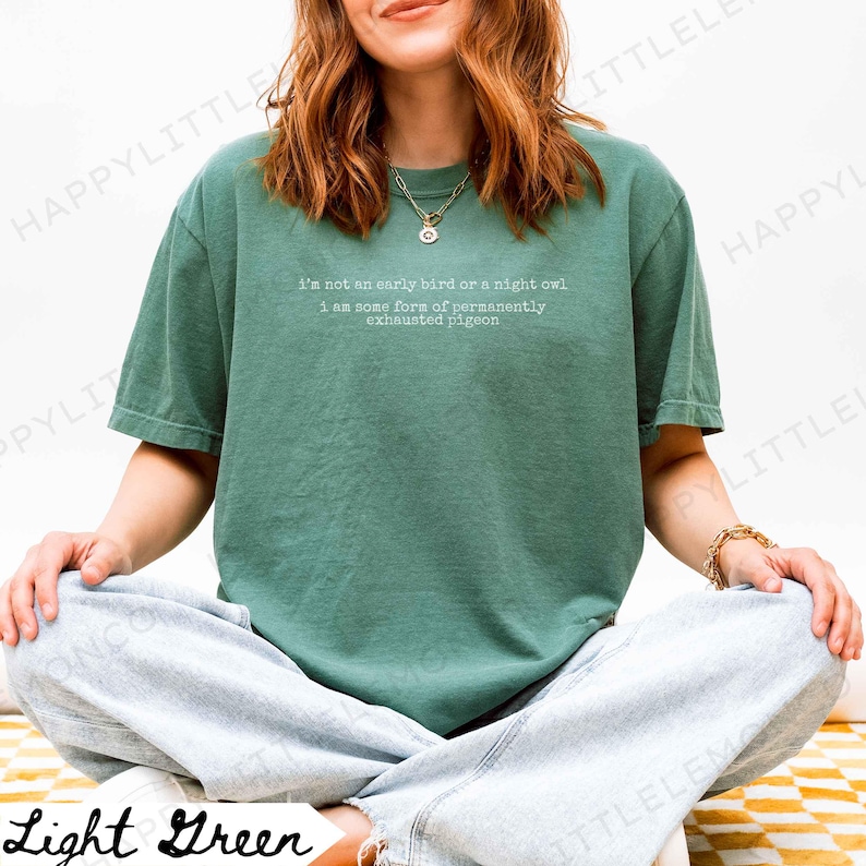 Puede incluir: Camiseta verde claro con el texto "I'm not an early bird or a night owl, I am some form of permanently exhausted pigeon." La camiseta es de color liso y tiene mangas cortas.