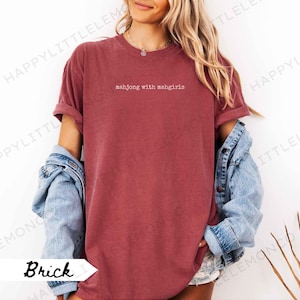 May include: Brick-coloured t-shirt with the text "mahjong with mahgirls" in white. The shirt has rolled-up sleeves and is paired with a denim jacket. The shirt is a casual, comfortable style.