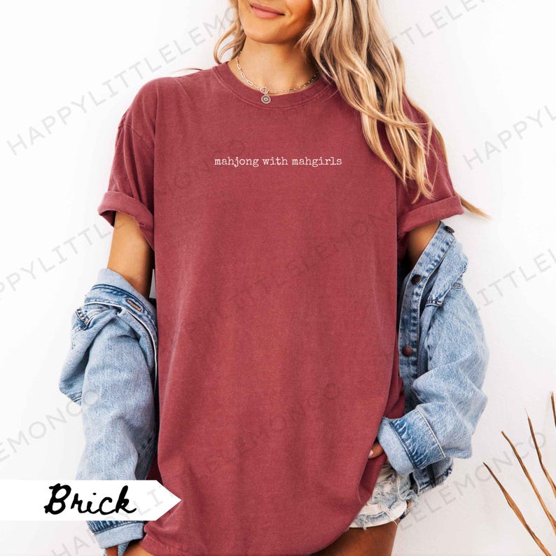 May include: Brick-coloured t-shirt with the text "mahjong with mahgirls" in white. The shirt has rolled-up sleeves and is paired with a denim jacket. The shirt is a casual, comfortable style.
