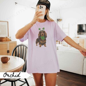 May include: Orchid-colored t-shirt featuring a dachshund wearing a green shamrock headband and scarf. The dog is brown and black. The shirt has a relaxed fit. A black baseball cap is worn.