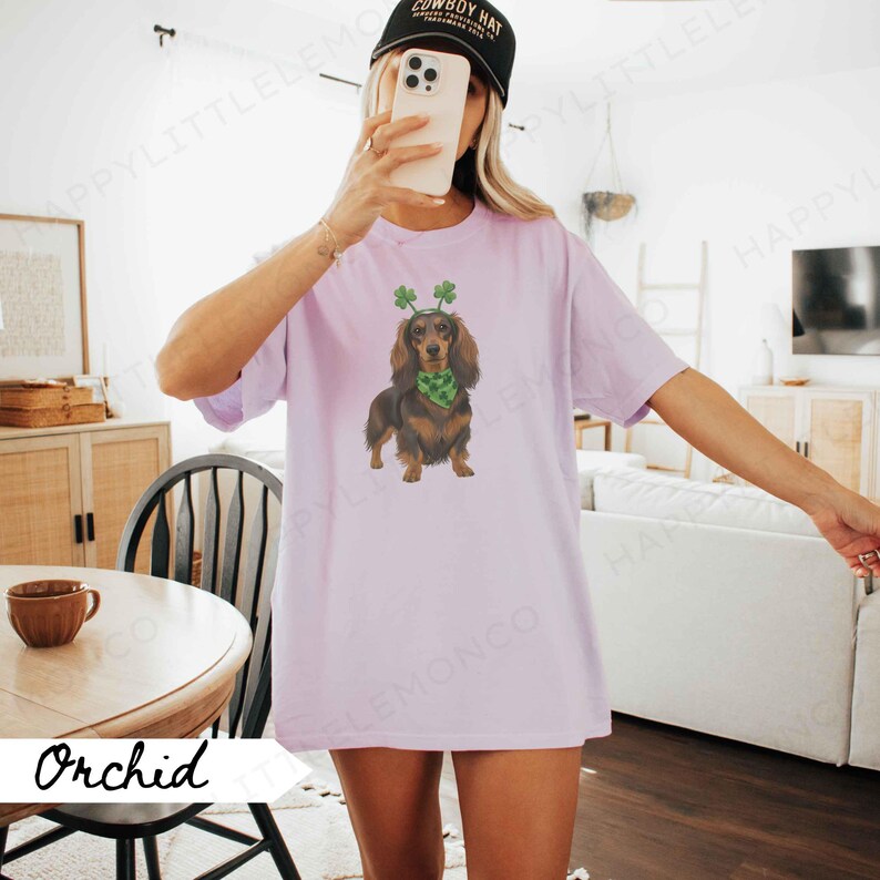 May include: Orchid-colored t-shirt featuring a dachshund wearing a green shamrock headband and scarf. The dog is brown and black. The shirt has a relaxed fit. A black baseball cap is worn.