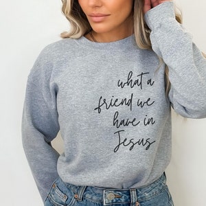 May include: A heather grey crewneck sweatshirt featuring the black cursive text 'what a friend we have in Jesus' printed on the front. The long-sleeved garment has a ribbed neckline, cuffs, and hem, providing a classic fit and casual appearance.