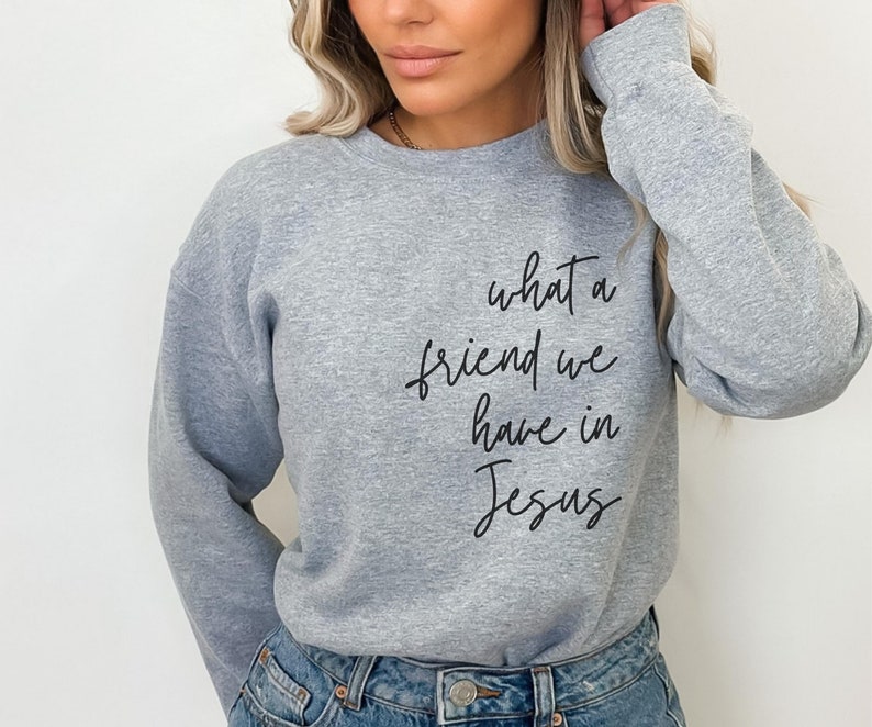 May include: A heather grey crewneck sweatshirt featuring the black cursive text 'what a friend we have in Jesus' printed on the front. The long-sleeved garment has a ribbed neckline, cuffs, and hem, providing a classic fit and casual appearance.