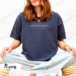 May include: Navy blue t-shirt featuring the white text "mahjong with mahgirls". The short-sleeved top has a relaxed fit. The person in the image is wearing light blue jeans and white trainers. The shirt is a casual style.