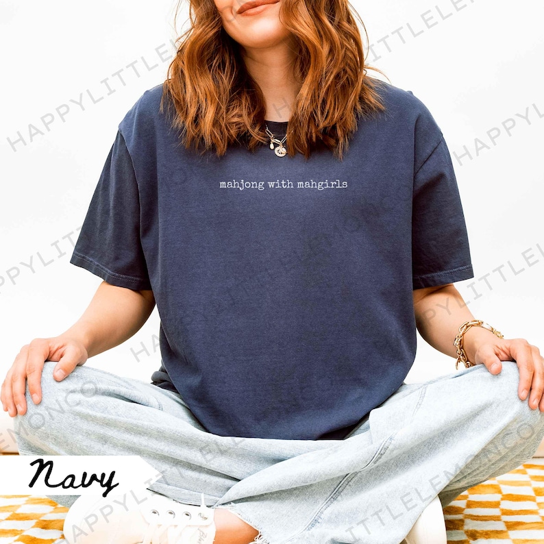 May include: Navy blue t-shirt featuring the white text "mahjong with mahgirls". The short-sleeved top has a relaxed fit. The person in the image is wearing light blue jeans and white trainers. The shirt is a casual style.