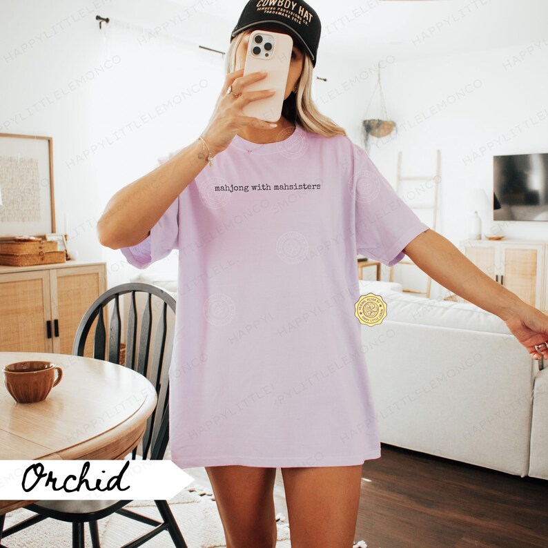 May include: A light purple t-shirt with the text "mahjong with mahsisters" in black. The shirt is worn with a black baseball cap. The word "Orchid" is written in white on a banner.