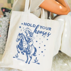 May include: A white canvas tote bag with blue text that reads "Hold Your Horses". The text is in a vintage style font. A cartoon image of a woman riding a horse is printed on the bag. The woman is wearing a cowboy hat and a dress. The horse is brown and white.