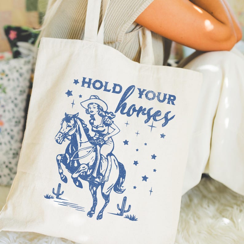 May include: A white canvas tote bag with blue text that reads "Hold Your Horses". The text is in a vintage style font. A cartoon image of a woman riding a horse is printed on the bag. The woman is wearing a cowboy hat and a dress. The horse is brown and white.