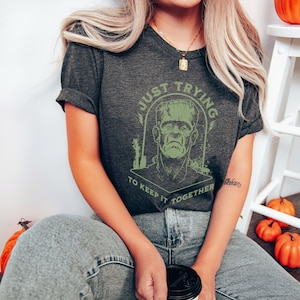 May include: A dark gray t-shirt with a green graphic of Frankenstein's monster and the text "Just Trying To Keep It Together".