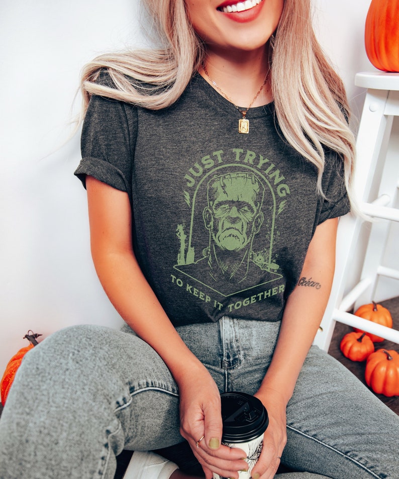 May include: A dark gray t-shirt with a green graphic of Frankenstein's monster and the text "Just Trying To Keep It Together".