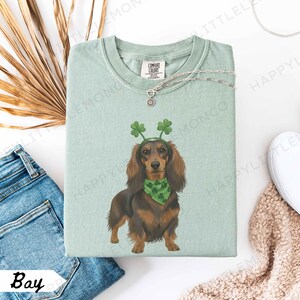 May include: A light green Comfort Colors t-shirt featuring a dachshund wearing a green shamrock headband and bandana. The dog is brown and black. A silver necklace and the word "Bay" are also visible.