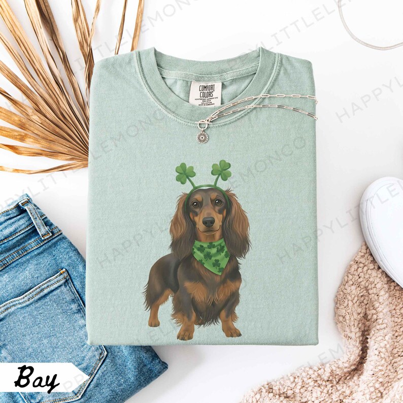 May include: A light green Comfort Colors t-shirt featuring a dachshund wearing a green shamrock headband and bandana. The dog is brown and black. A silver necklace and the word "Bay" are also visible.