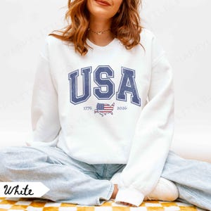 May include: White sweatshirt with the word "USA" in blue block letters. Below the word "USA" is the text "1776" and "2026" with a graphic of the American flag in the shape of the United States. The sweatshirt is a crew neck.