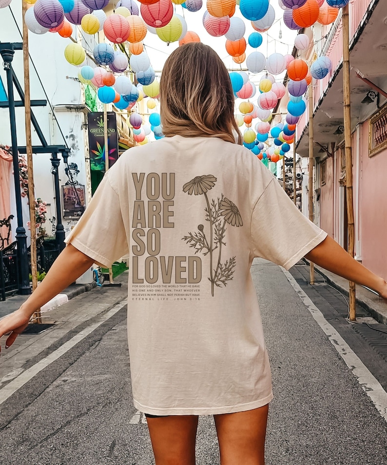 May include: A beige t-shirt with the words "YOU ARE SO LOVED" printed on the back, along with a floral design and a Bible verse. The shirt is a relaxed fit. The image is taken outdoors with colorful paper lanterns in the background.