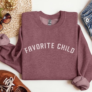May include: A mauve heather crewneck sweatshirt featuring the text FAVORITE CHILD printed in a distressed white font across the chest. The garment has long sleeves with ribbed cuffs and a ribbed neckline, showcasing a soft, textured fabric finish.