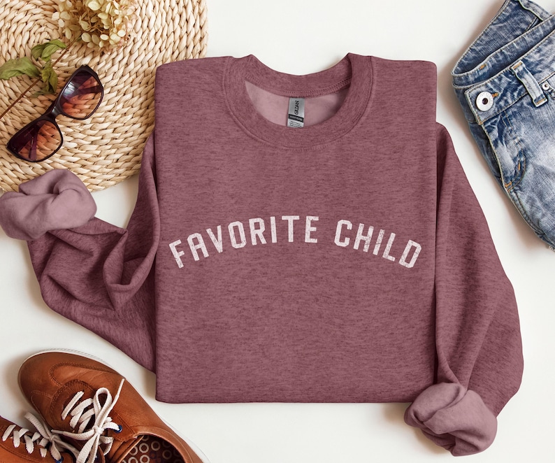 May include: A mauve heather crewneck sweatshirt featuring the text FAVORITE CHILD printed in a distressed white font across the chest. The garment has long sleeves with ribbed cuffs and a ribbed neckline, showcasing a soft, textured fabric finish.