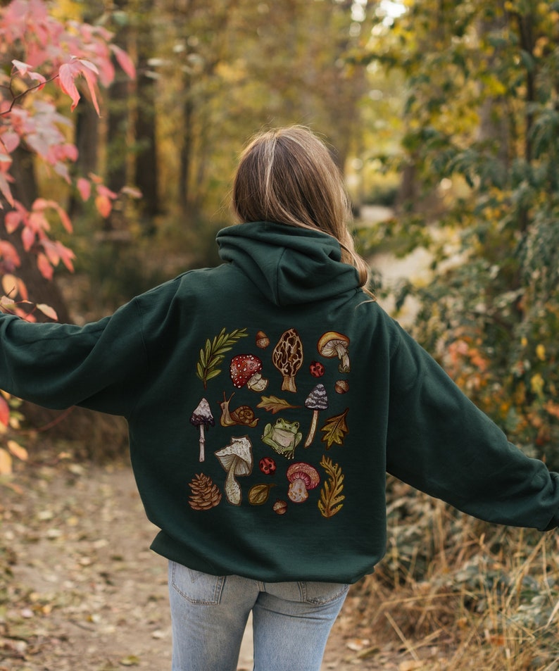 May include: A dark green hooded sweatshirt with a colorful mushroom and frog design on the back. The design features various mushrooms, a frog, a snail, leaves, and pine cones.