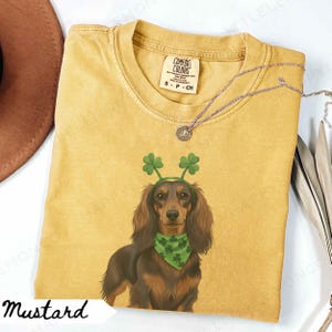May include: Mustard-colored t-shirt featuring a dachshund wearing a green shamrock headband and bandana. The shirt has a Comfort Colors tag and a small silver necklace. The word "Mustard" is written in a script font at the bottom.