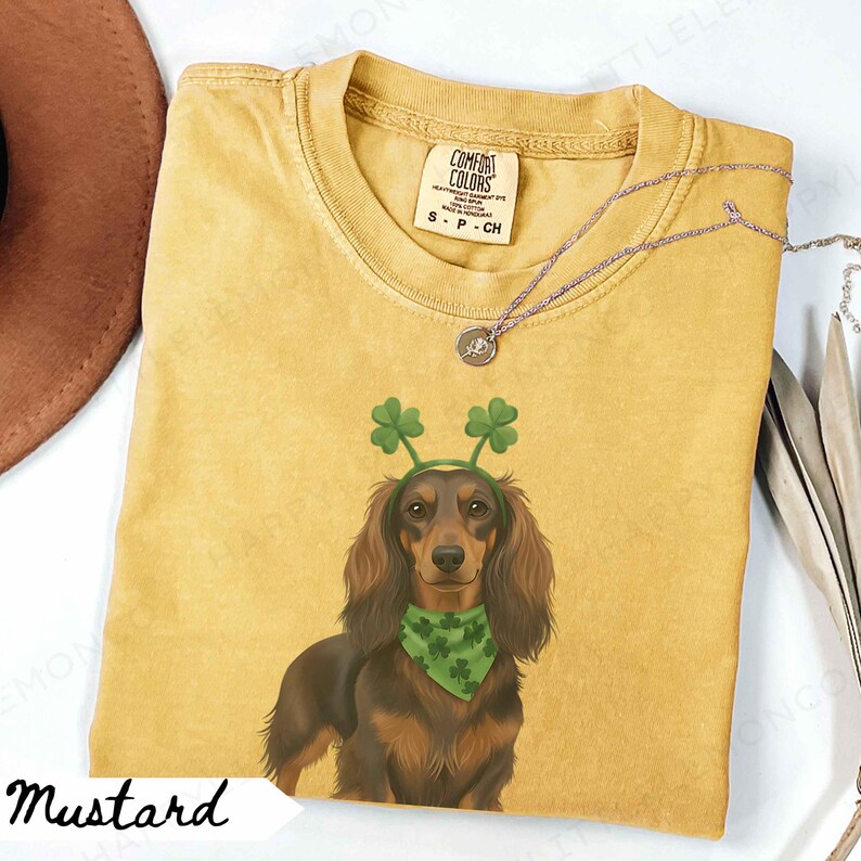 May include: Mustard-colored t-shirt featuring a dachshund wearing a green shamrock headband and bandana. The shirt has a Comfort Colors tag and a small silver necklace. The word "Mustard" is written in a script font at the bottom.