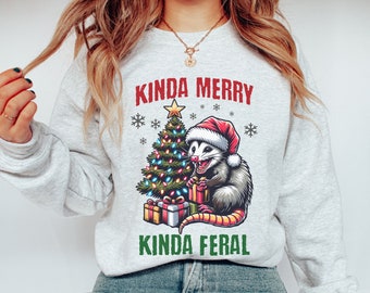 May include: A light gray sweatshirt with a graphic featuring a possum wearing a Santa hat, holding gifts next to a Christmas tree. The text reads "Kinda Merry Kinda Feral" in red and green.