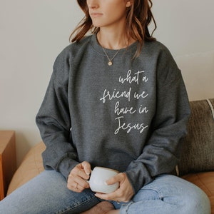 May include: A dark charcoal grey crewneck sweatshirt featuring the white cursive text "what a friend we have in Jesus" printed on the upper right chest area. The long-sleeved garment has a ribbed neckline, cuffs, and hem, with a simple design.