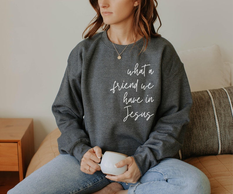 May include: A dark charcoal grey crewneck sweatshirt featuring the white cursive text "what a friend we have in Jesus" printed on the upper right chest area. The long-sleeved garment has a ribbed neckline, cuffs, and hem, with a simple design.