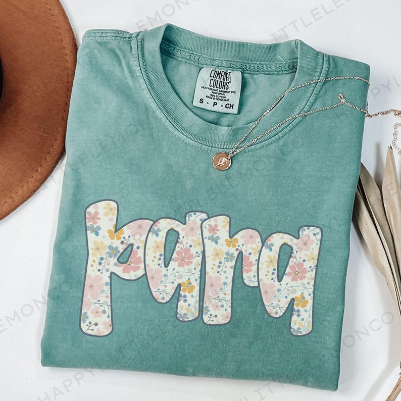 May include: A teal Comfort Colors t-shirt with the word "mama" in a floral design. The letters are outlined in white and filled with pink, yellow, and blue flowers. A gold necklace with a pendant is draped across the shirt.