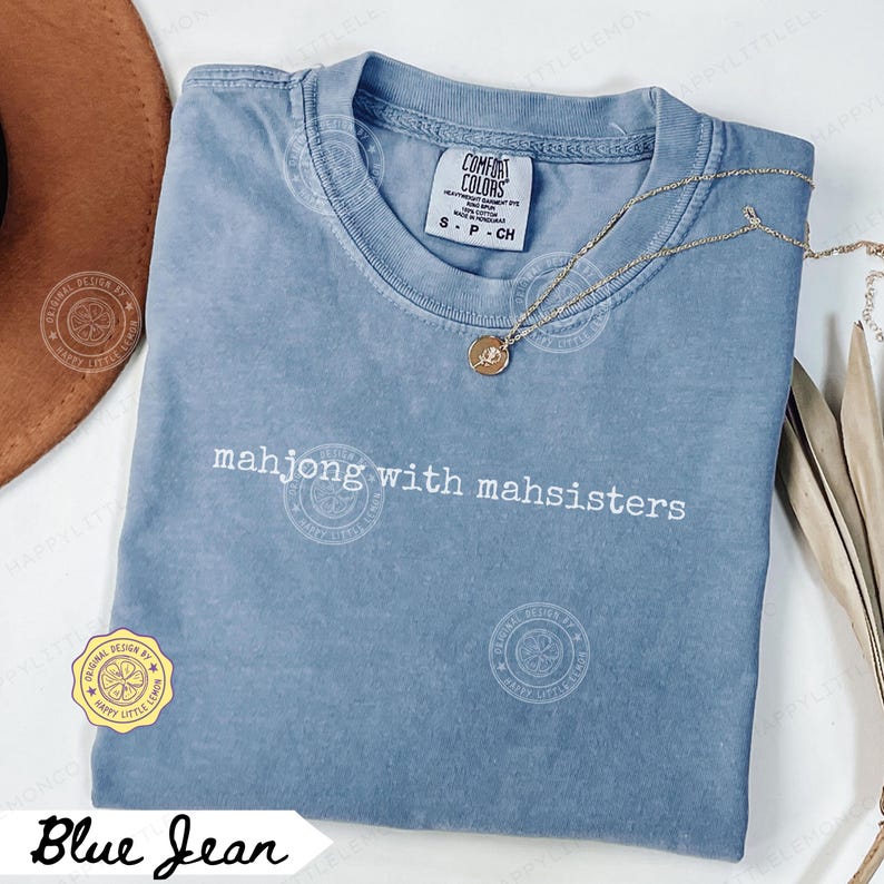 May include: A blue jean colored t-shirt with the text "mahjong with mahsisters" in white. The shirt has a crew neck and short sleeves. A gold necklace with a pendant is draped across the shirt. The shirt is folded on a white surface.
