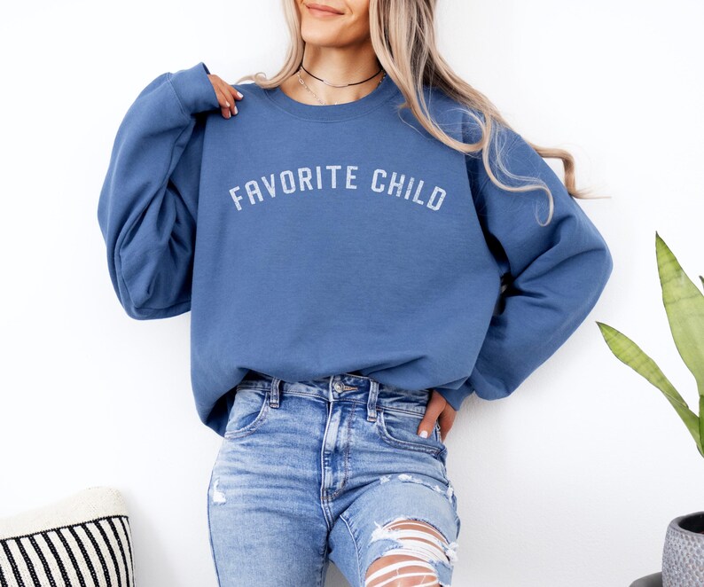 May include: A blue crewneck sweatshirt featuring the text FAVORITE CHILD printed in a white, slightly distressed, arched font across the chest. The garment has long sleeves and a ribbed neckline, cuffs, and hem, paired with light blue distressed denim jeans.