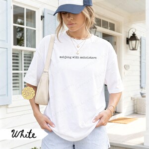 May include: White t-shirt with the text "mahjong with mahsisters" in a black, simple font. The shirt is worn with a blue and white baseball cap and striped shorts. A cream-colored purse is worn over the shoulder.