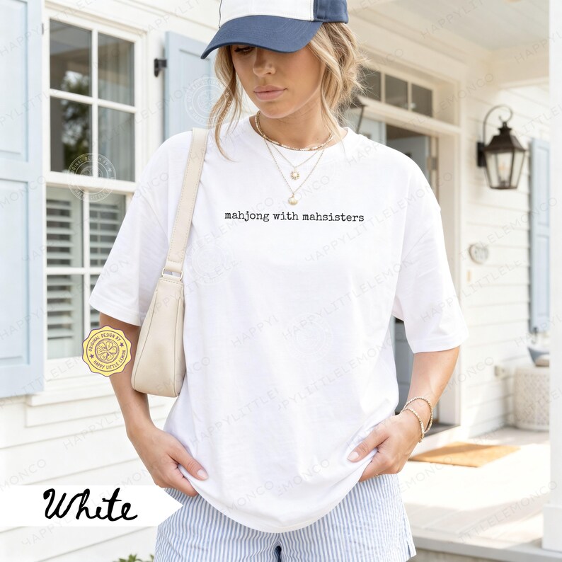 May include: White t-shirt with the text "mahjong with mahsisters" in a black, simple font. The shirt is worn with a blue and white baseball cap and striped shorts. A cream-colored purse is worn over the shoulder.