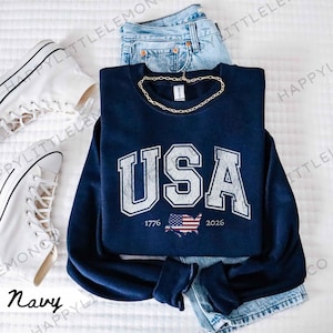 May include: Navy blue sweatshirt with "USA" in distressed white lettering, the years 1776 and 2026, and a red, white, and blue American flag graphic. The sweatshirt is paired with blue jeans, a gold chain, and white sneakers.