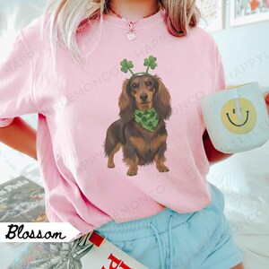 May include: Pink Blossom sweatshirt featuring a dachshund wearing a green bandana and shamrock headband. The dog illustration is brown and black. A white mug with a yellow smiley face is in the background.