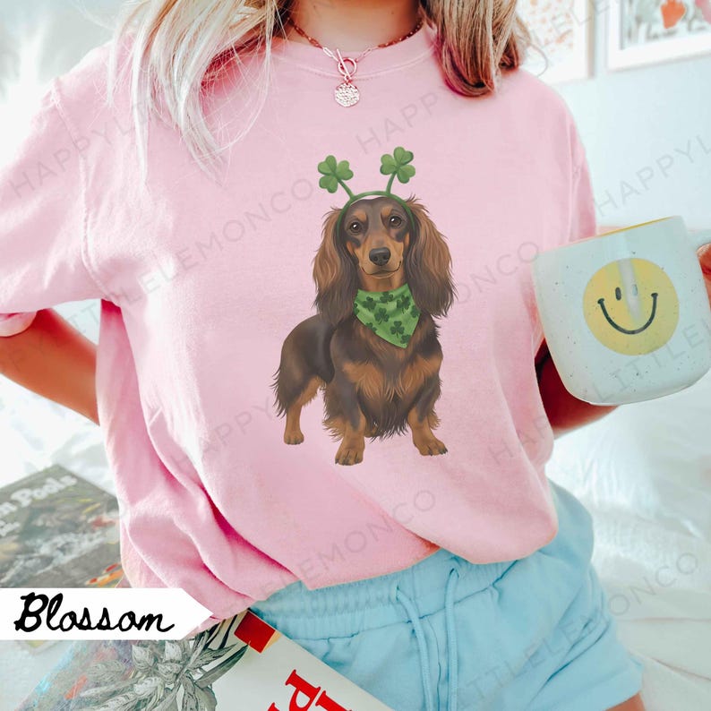 May include: Pink Blossom sweatshirt featuring a dachshund wearing a green bandana and shamrock headband. The dog illustration is brown and black. A white mug with a yellow smiley face is in the background.