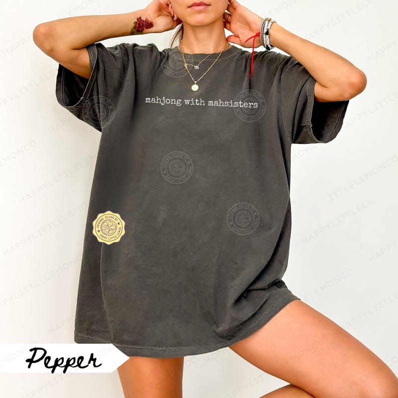 May include: A dark gray t-shirt with the text "mahjong with mahsisters" in white. The shirt has a relaxed fit and a vintage look. A small gold sticker is on the lower left side of the shirt. The word "Pepper" is written in cursive at the bottom.