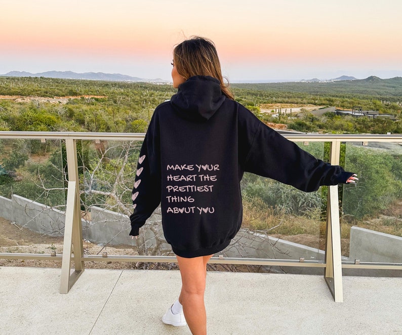 Female model wearing a black hoodie.  The back of the hoodie has the quote 'Make Your Heart The Prettiest Thing About You' printed on the back lower half. The left sleeve has 5 hearts printed on it.