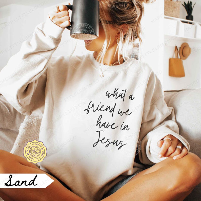 May include: A sand-coloured crewneck sweatshirt featuring the black cursive text 'what a friend we have in Jesus' printed on the front. The garment has long sleeves and a ribbed neckline, cuffs, and hem, providing a relaxed fit for casual wear.
