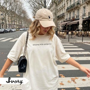May include: An ivory-coloured t-shirt with the text "mahjong with mahgirls" in black. A beige baseball cap with a black logo is worn. A black quilted shoulder bag with a chain strap is also visible.