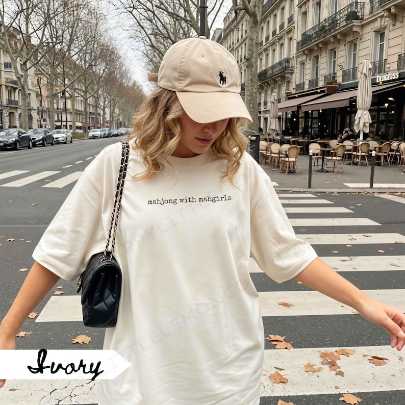 May include: An ivory-coloured t-shirt with the text "mahjong with mahgirls" in black. A beige baseball cap with a black logo is worn. A black quilted shoulder bag with a chain strap is also visible.