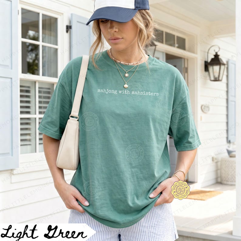 May include: Light green t-shirt with the text "mahjong with mahsisters" in white. The shirt is a relaxed fit and is paired with striped shorts. A cream-colored purse is slung over the shoulder.