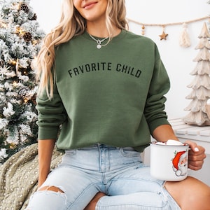 May include: An olive green crewneck sweatshirt featuring the text FAVORITE CHILD printed in black across the chest. The garment has long sleeves and a ribbed neckline, cuffs, and hem, providing a standard fit for casual wear during the holiday season.