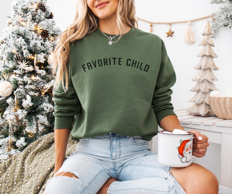 May include: An olive green crewneck sweatshirt featuring the text FAVORITE CHILD printed in black across the chest. The garment has long sleeves and a ribbed neckline, cuffs, and hem, providing a standard fit for casual wear during the holiday season.